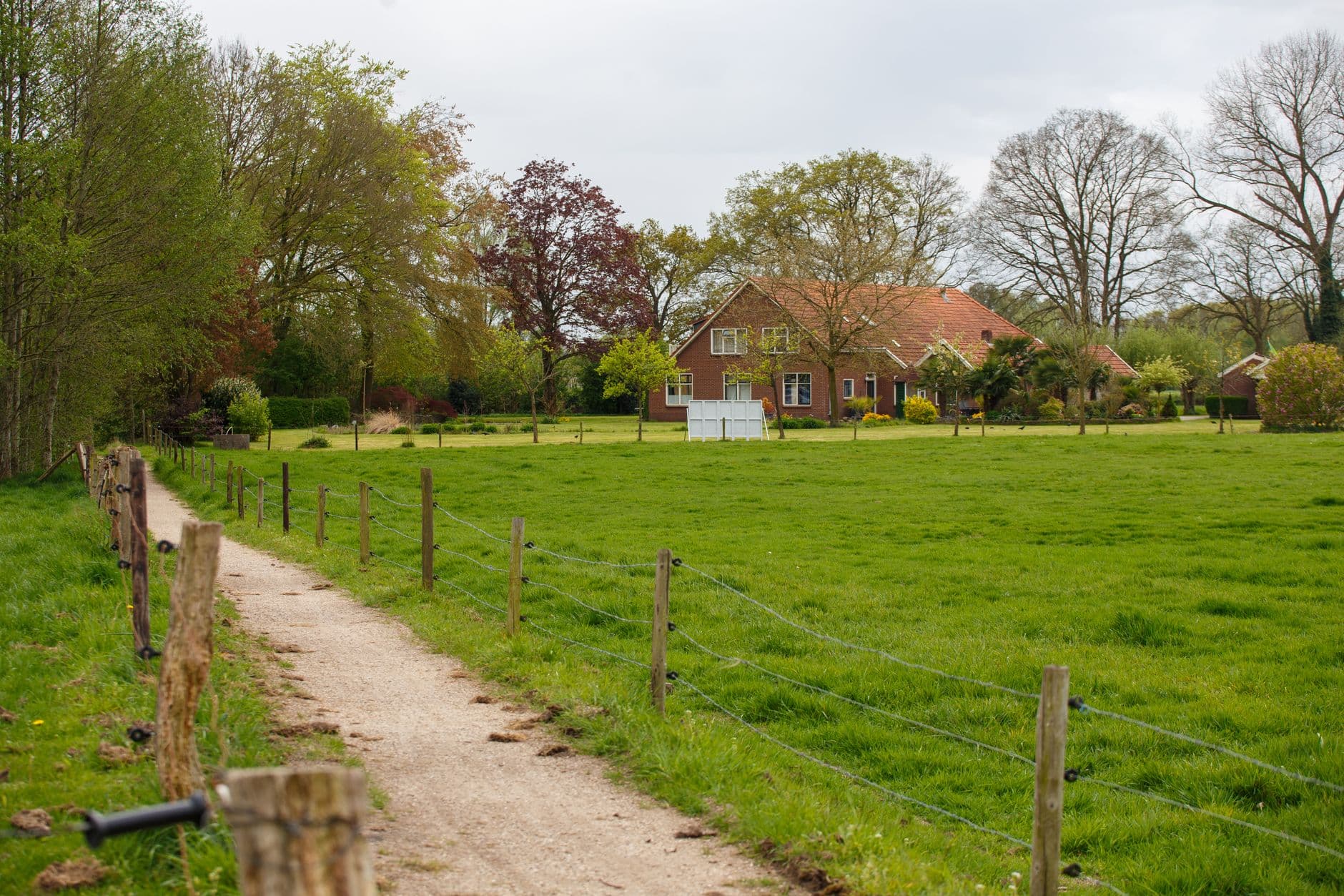 Rode bakstenen woning achter een groene weide met houten afrastering en een onverhard pad