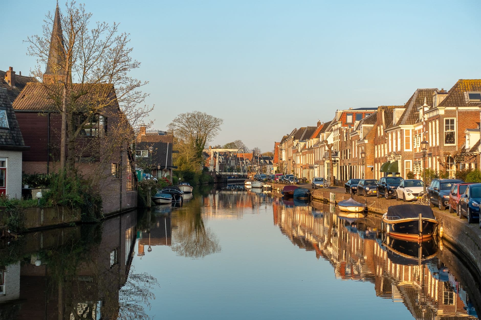 Bakstenen huizen, geparkeerde auto's en aangemeerde bootjes langs een kanaal in laag zonlicht