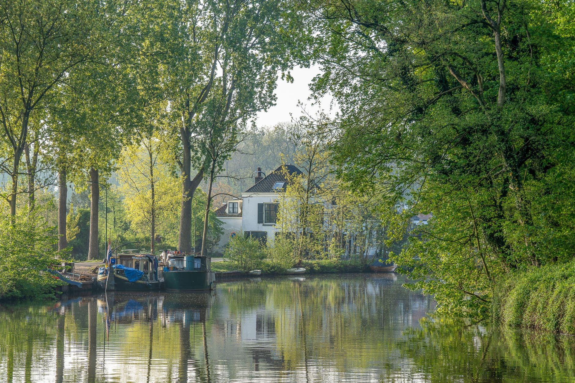Stille waterloop met aangemeerde boten en een wit huis tussen bomen, weerspiegeld in het water