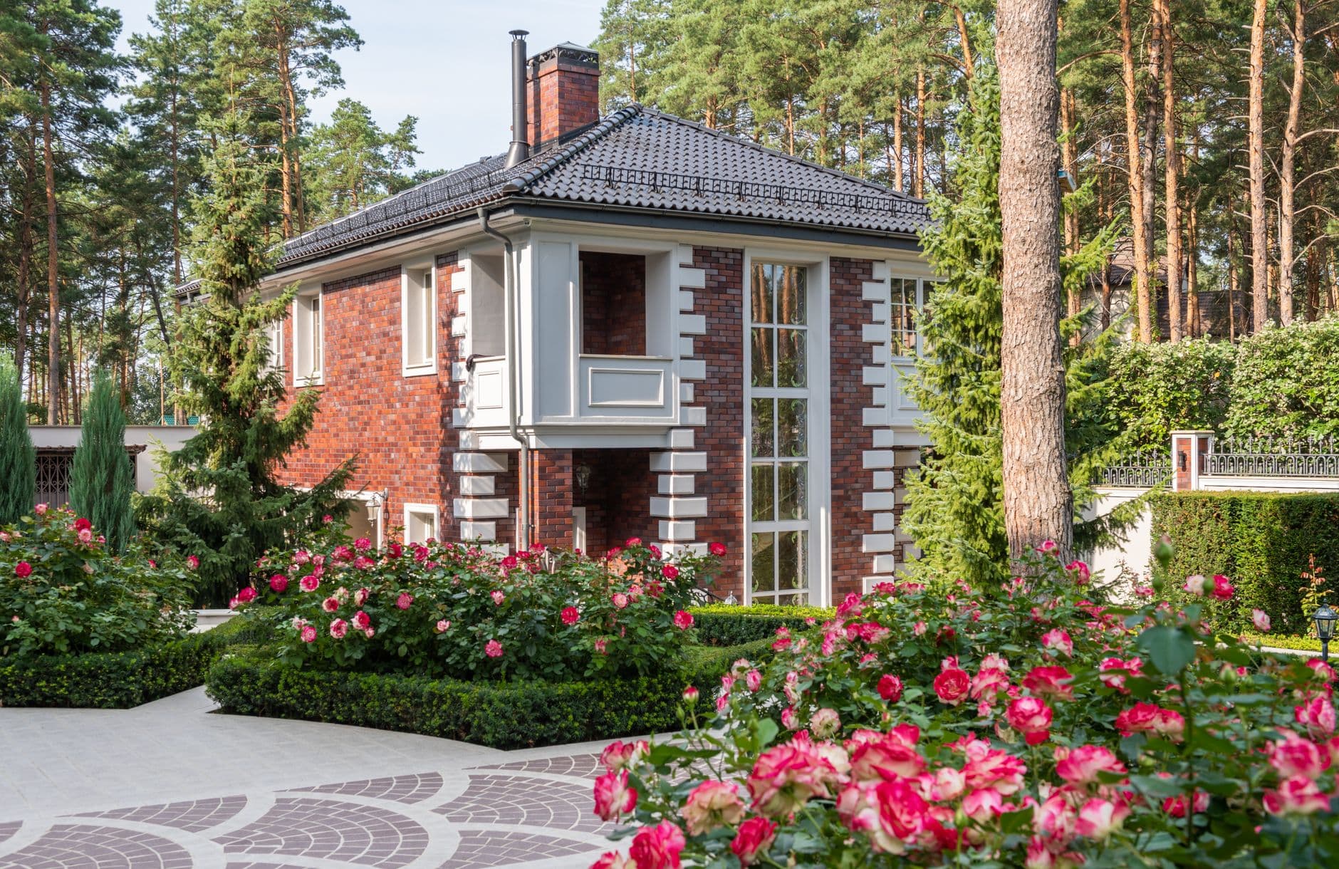 Bakstenen villa met donker pannendak tussen hoge dennen, met bloeiende rozen in de voortuin