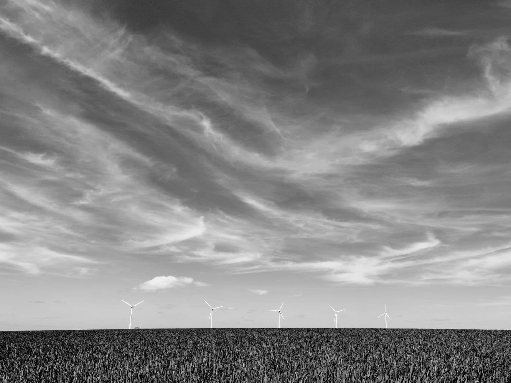 Zwart-witbeeld van windturbines aan de horizon achter een akker onder een wolkenlucht