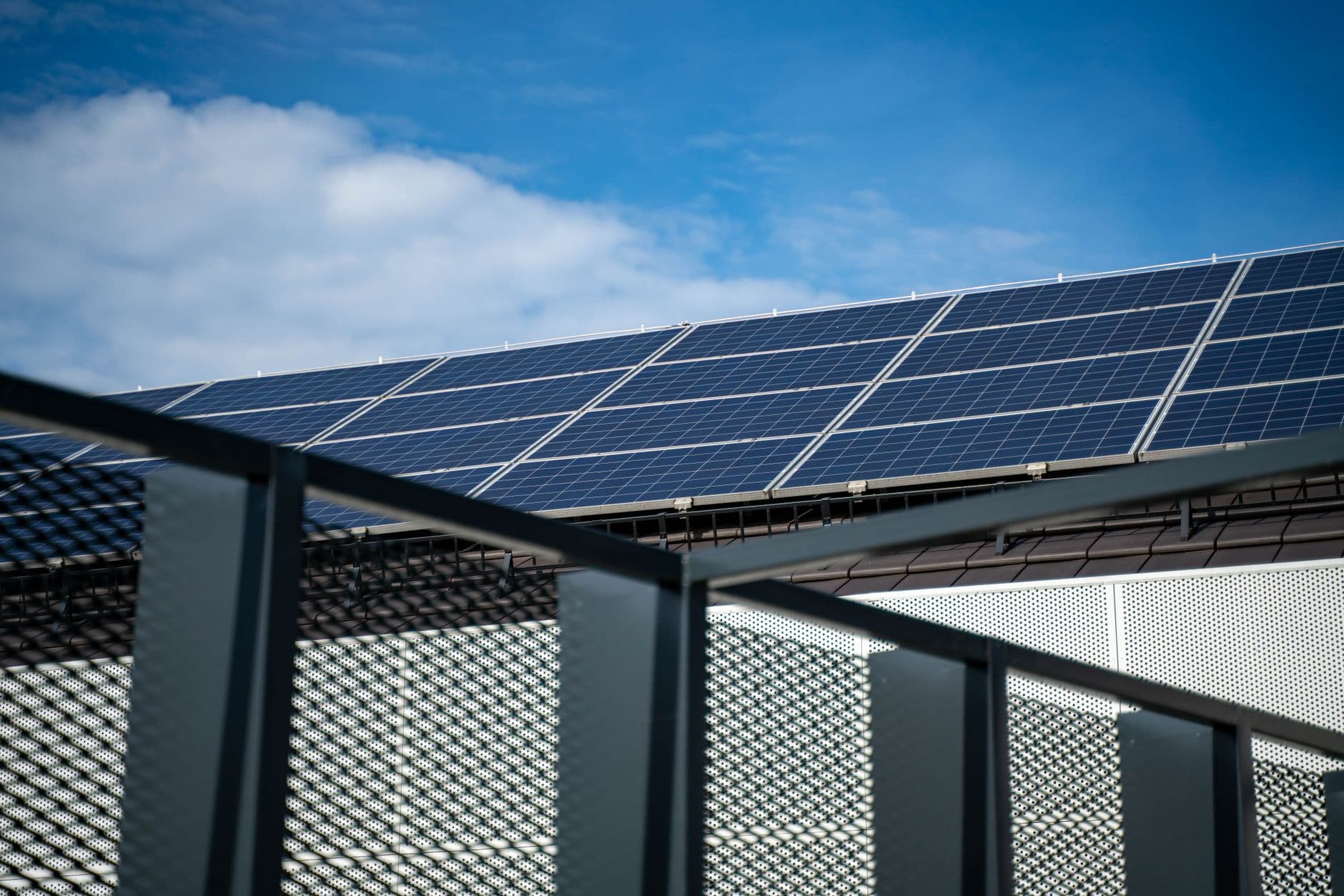 Zonnepanelen op een dak achter een metalen balustrade, onder een blauwe lucht met wolken