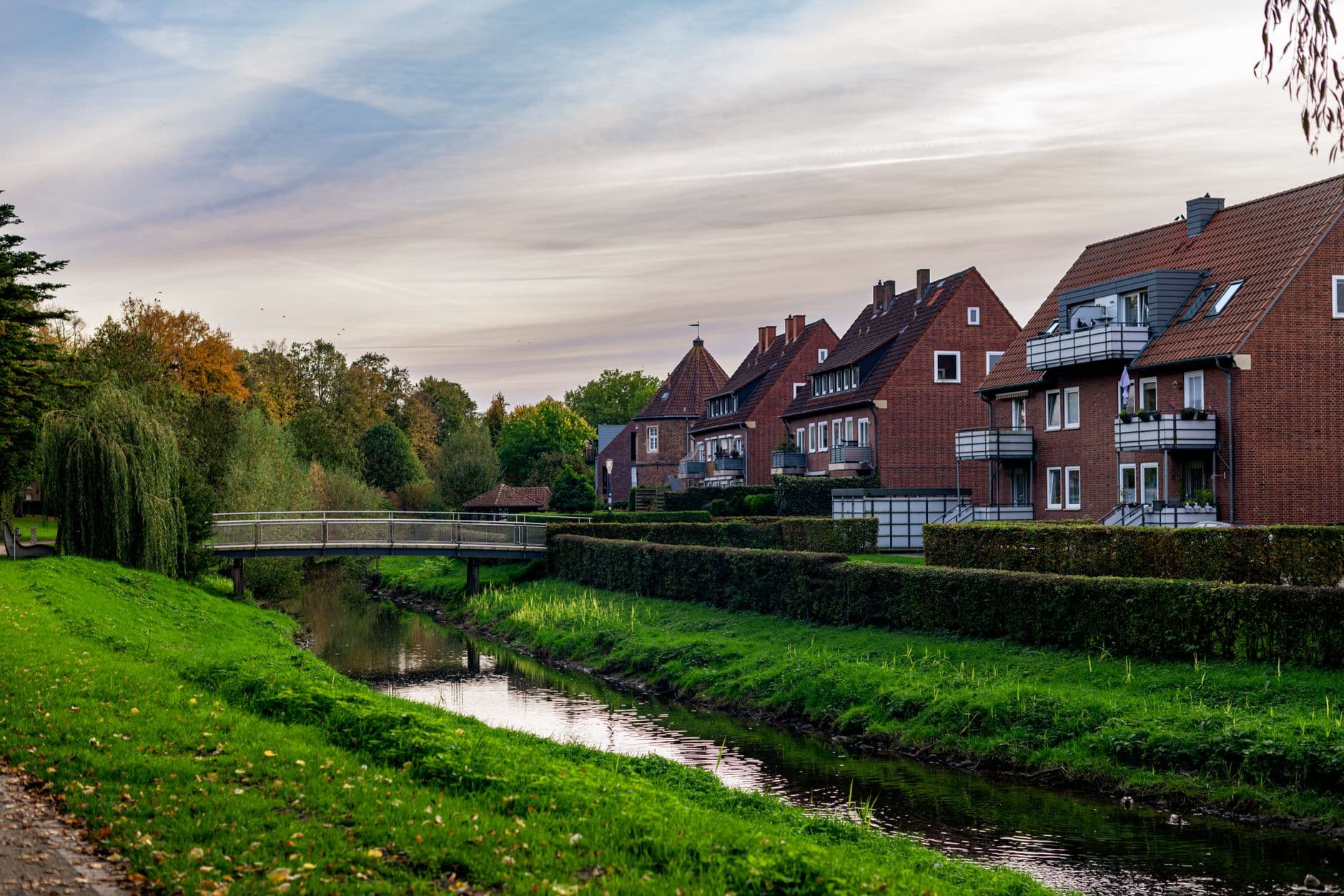 Rode bakstenen woningen langs een beek met een voetgangersbrug en herfstbomen