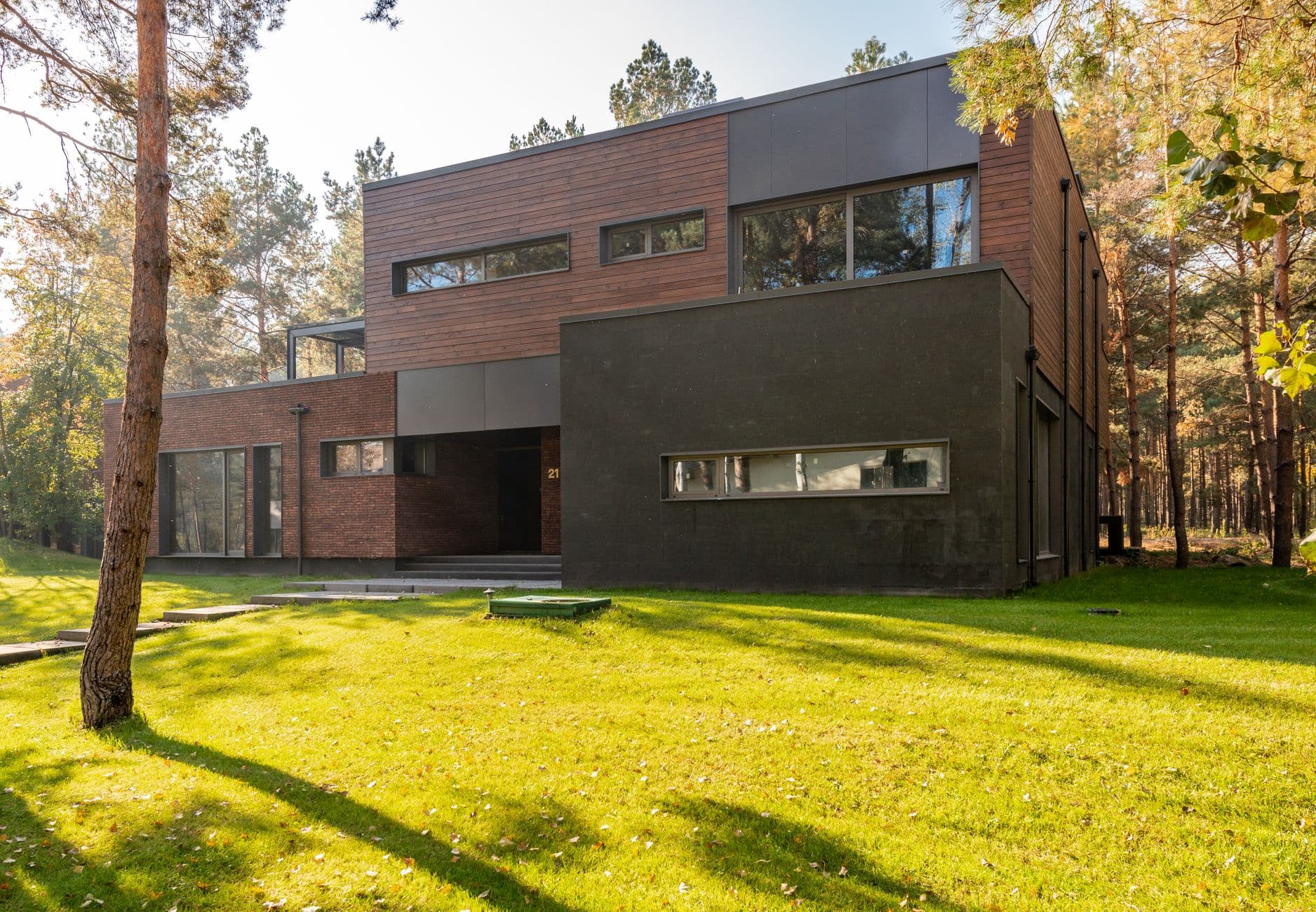 Moderne woning met bakstenen gevel en donkere volumes op een zonnig gazon tussen dennenbomen