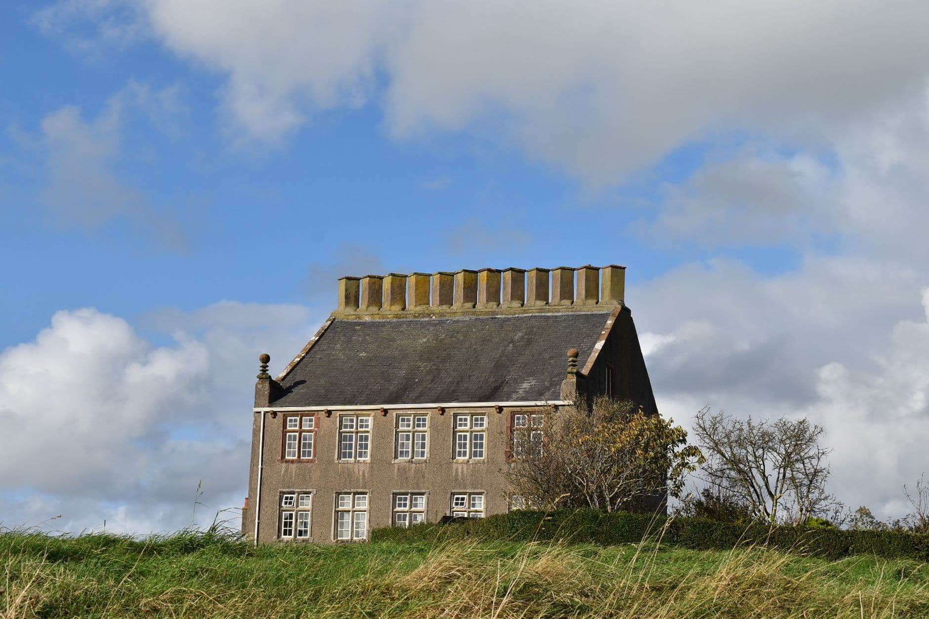 Oud bakstenen huis met leien dak op een groene heuvel onder een blauwe lucht met wolken