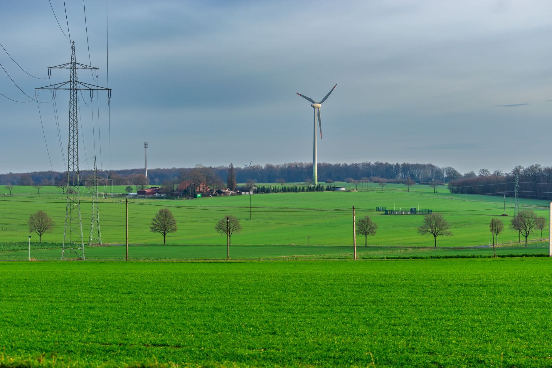 Groene akkers met een windturbine, hoogspanningsmasten en boerderijen onder een grijze lucht