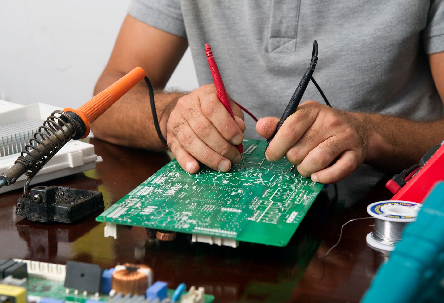 Handen van een technicus die met meetpennen een printplaat controleren naast een soldeerbout