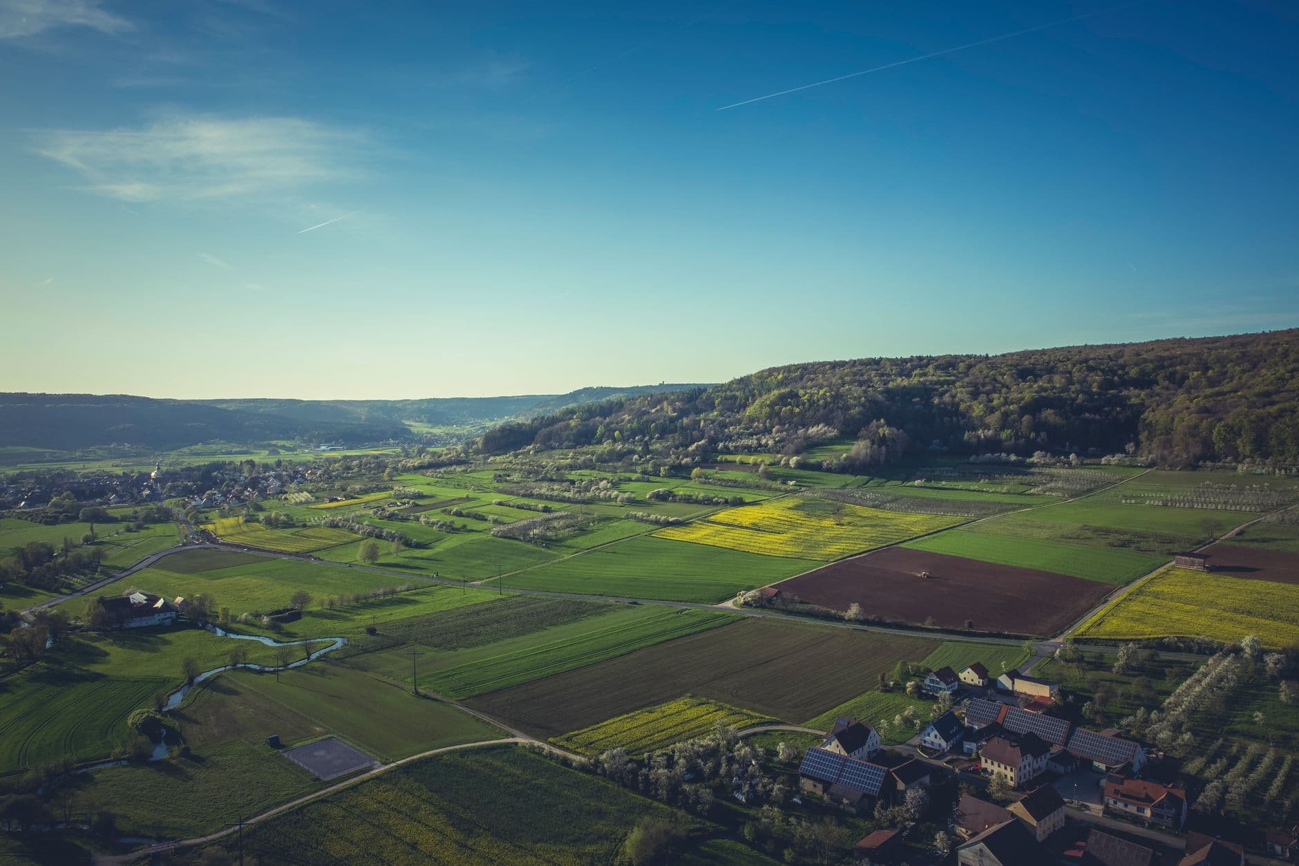 Luchtbeeld van akkers en weiden rond een dorp, enkele daken met zonnepanelen en beboste heuvels