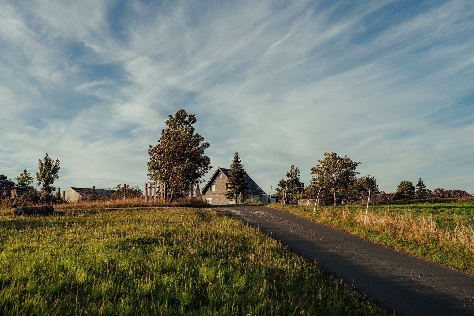 Landelijke weg door vlak grasland naar een boerderij tussen bomen onder een wijde wolkenlucht