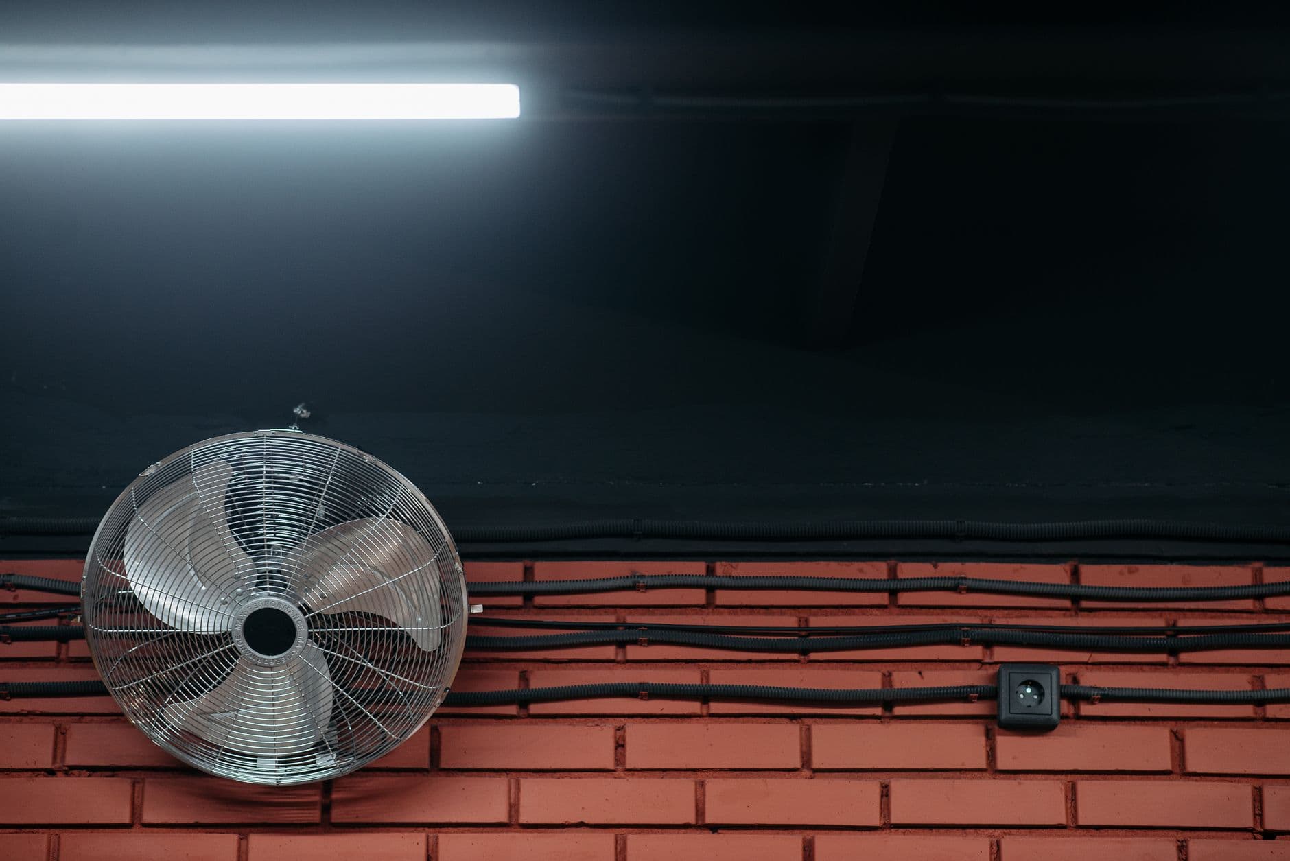 Ventilator, opbouwbekabeling en een stopcontact tegen een rode bakstenen muur onder een tl-lamp