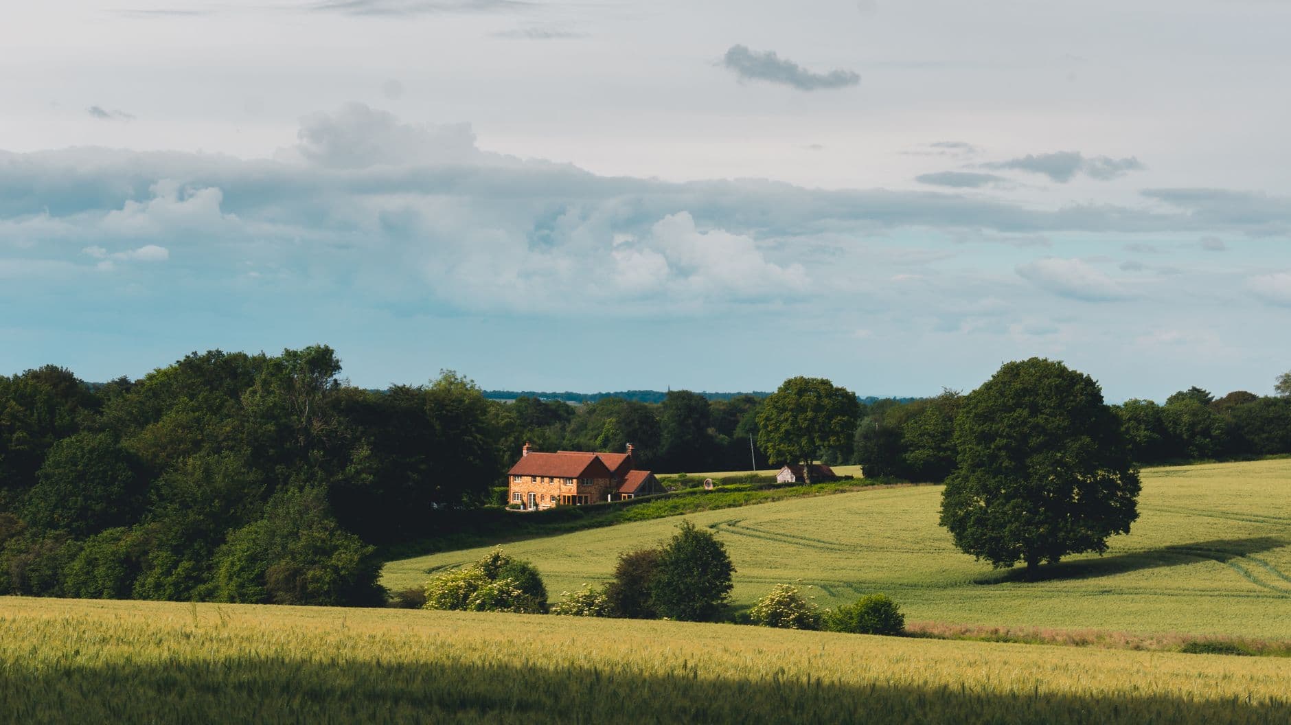 Bakstenen landhuis met rode pannen tussen bomen en golvende graanvelden onder een bewolkte lucht