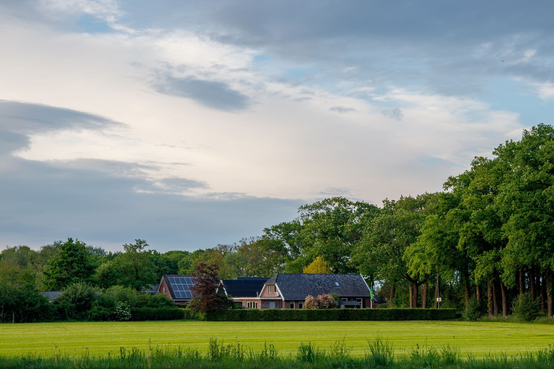 Bakstenen hoeve met zonnepanelen op het dak achter een haag en een groen grasveld