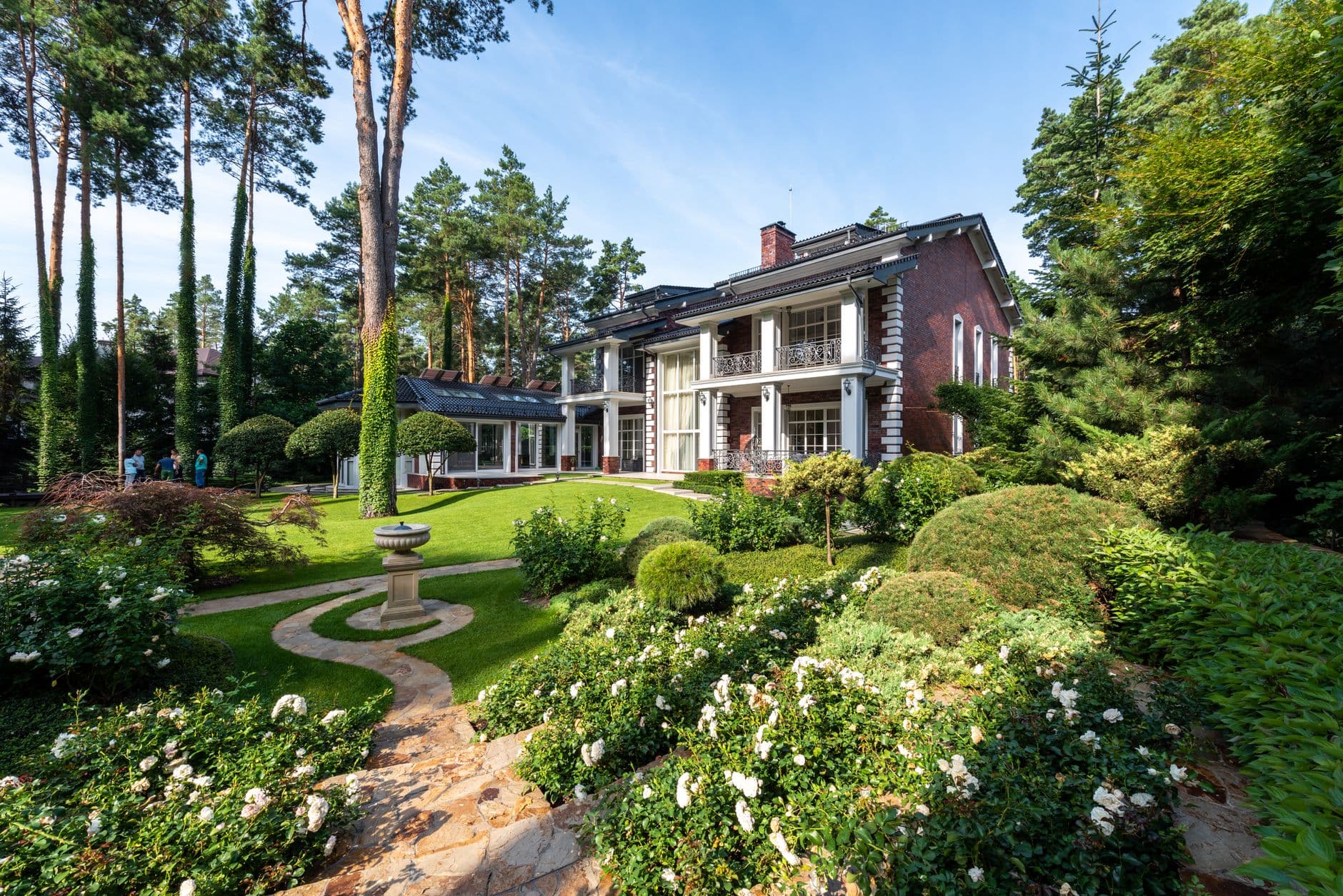 Bakstenen villa met witte zuilen tussen hoge dennen en een tuin met bloeiende struiken
