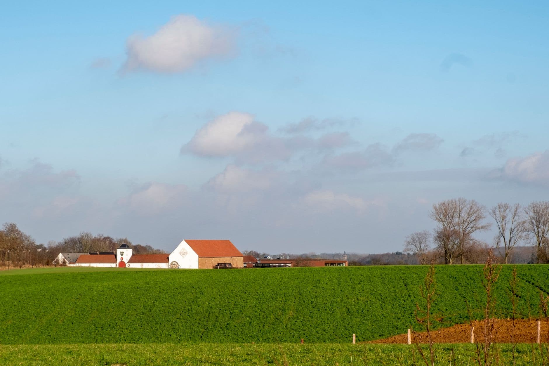 Witte vierkantshoeve met rode daken achter een groene akker onder een blauwe lucht
