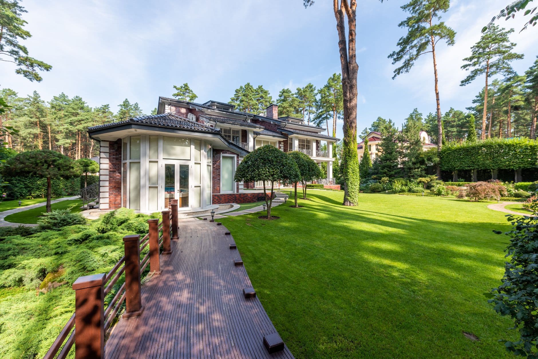 Ruime villa met deels bakstenen gevel, houten terras en verzorgd gazon tussen hoge dennen