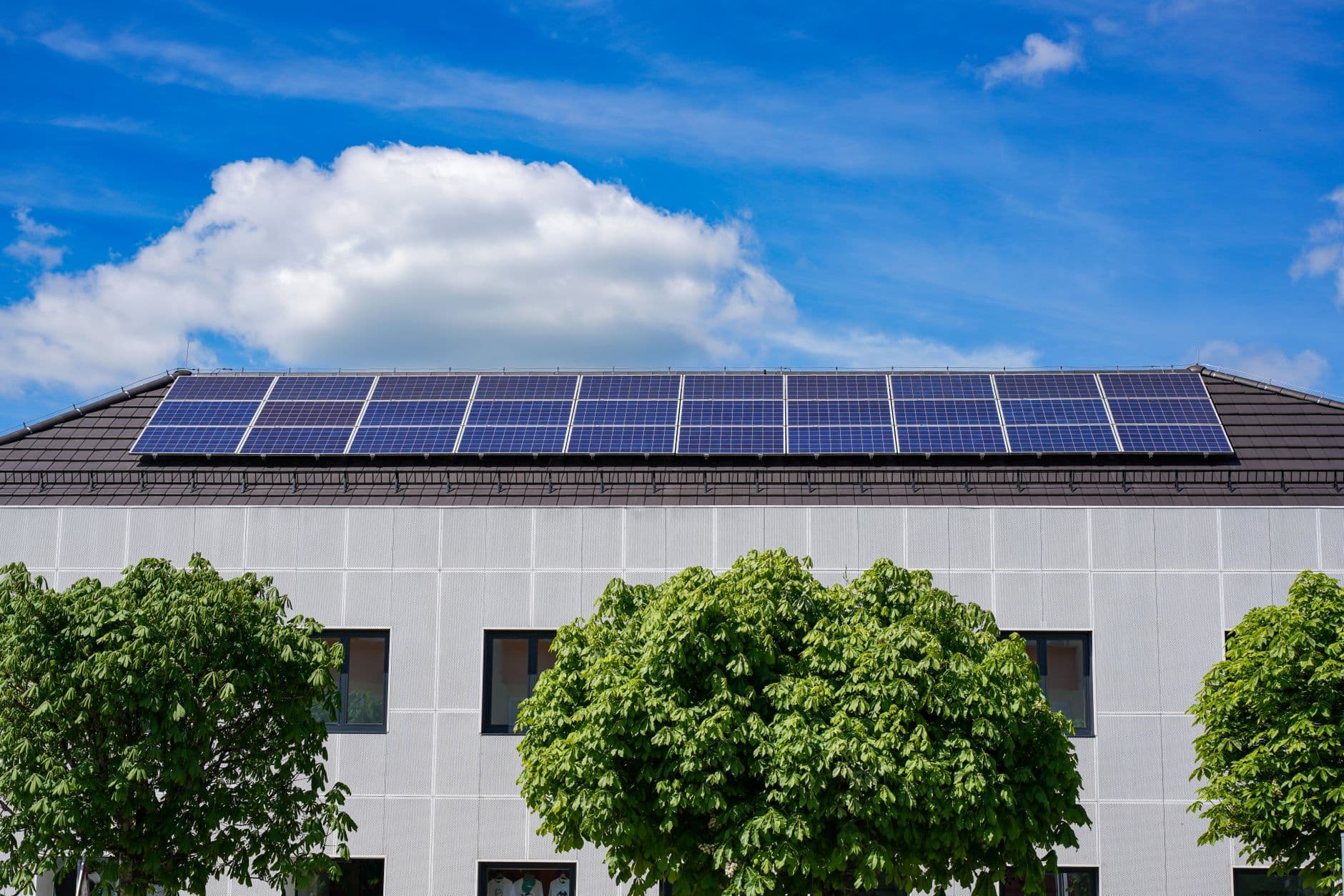 Zonnepanelen op het dak van een gebouw met lichte gevel, groene bomen ervoor en een blauwe lucht