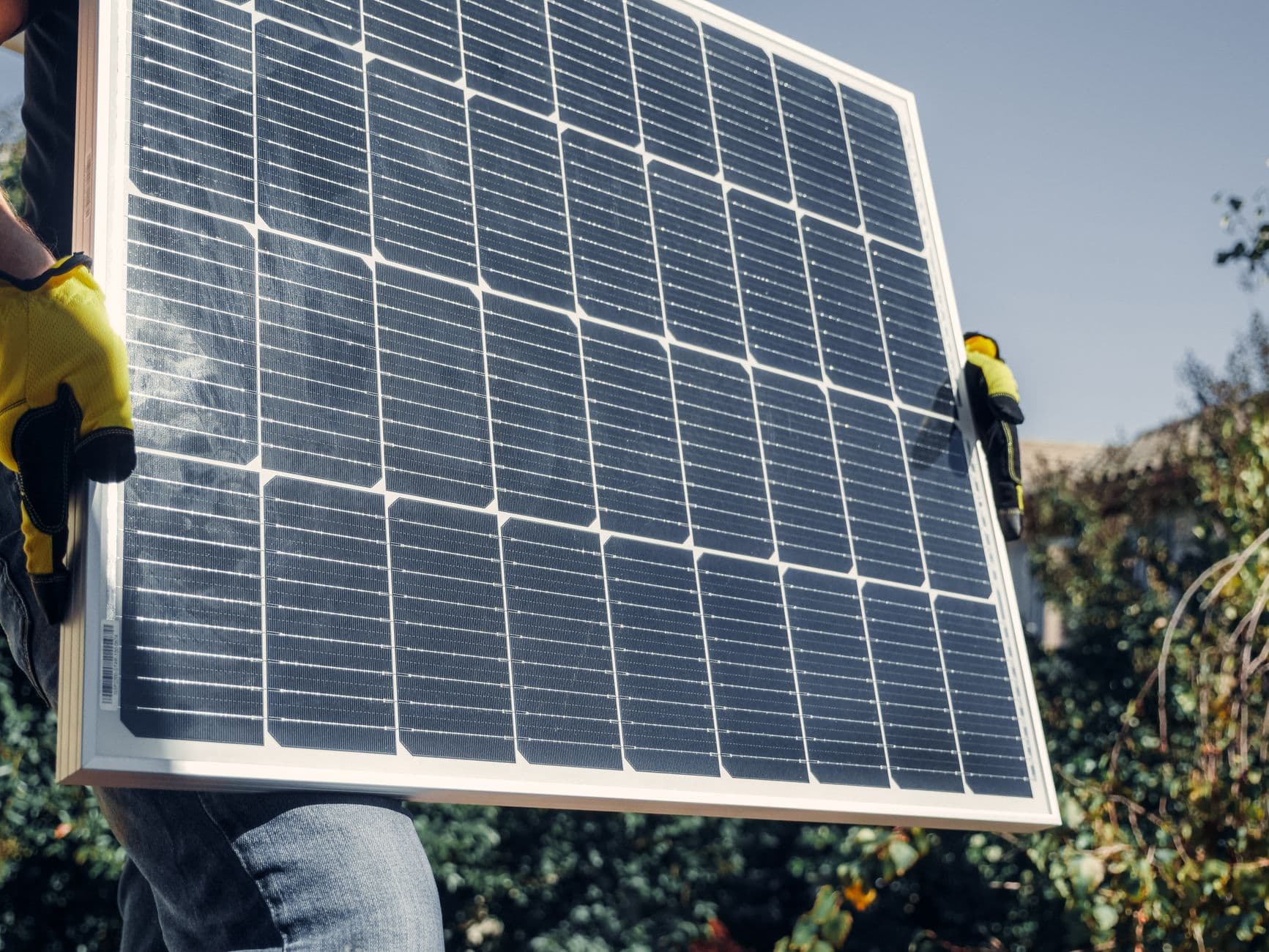 Handen in werkhandschoenen dragen een zonnepaneel buiten tussen het groen