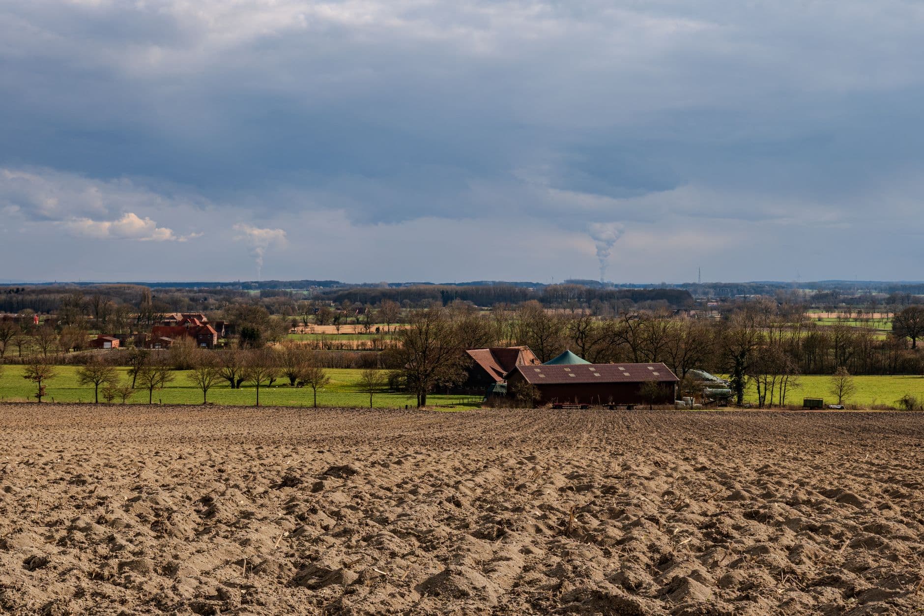 Omgeploegde akker met boerderijgebouwen, bomen en groene weiden onder een bewolkte lucht