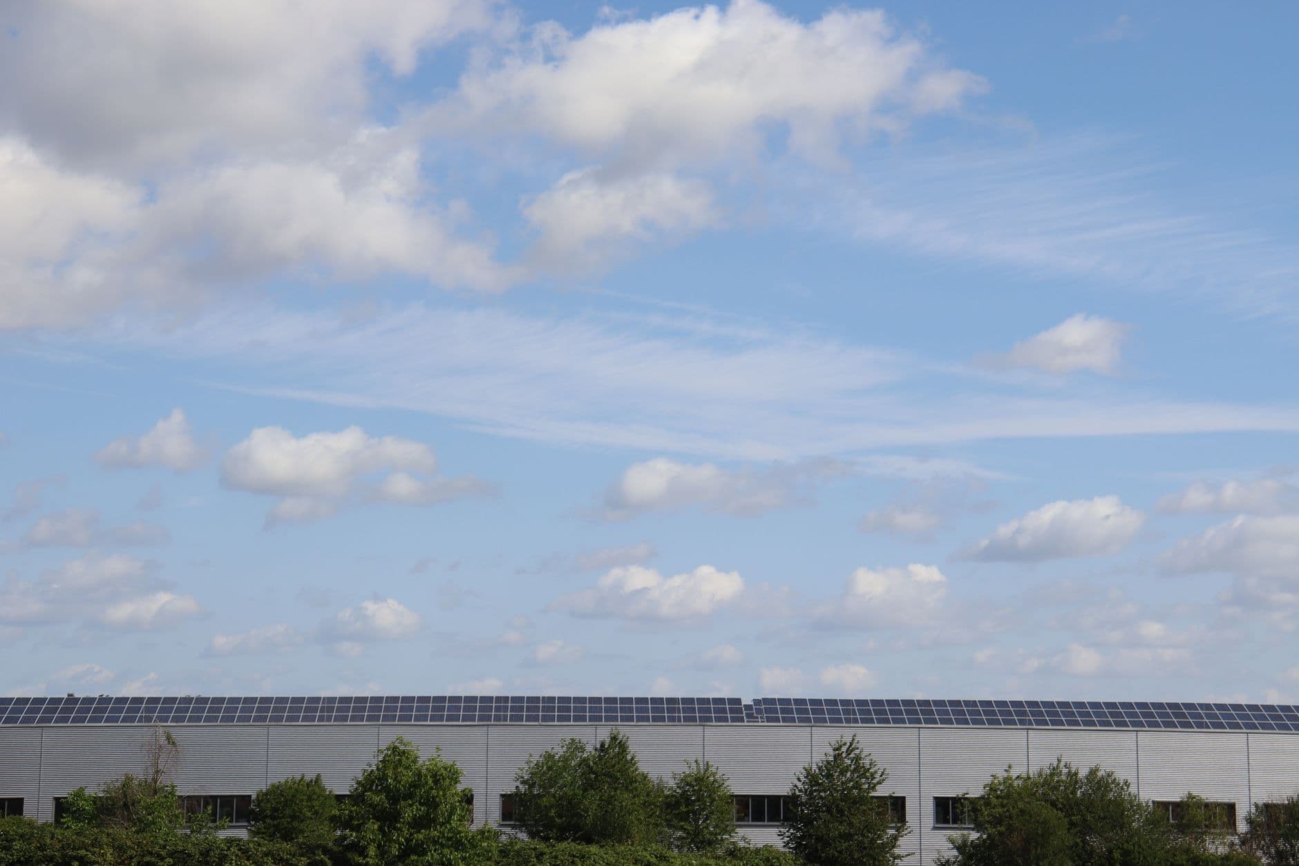 Bedrijfshal met zonnepanelen op het dak en bomen ervoor onder een blauwe lucht met wolken