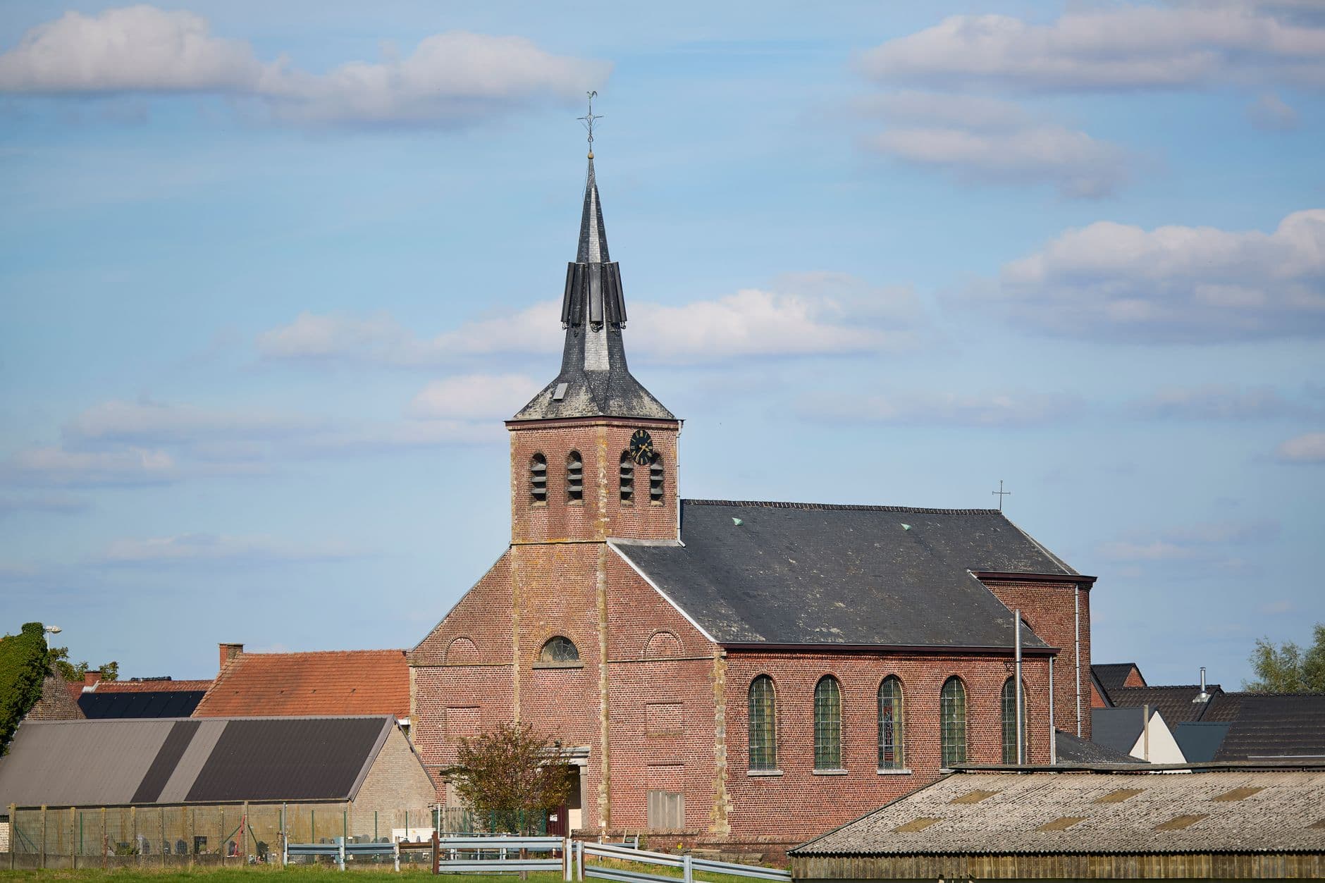 Bakstenen dorpskerk met leien torenspits onder een blauwe lucht, met zonnepanelen op een dak ernaast