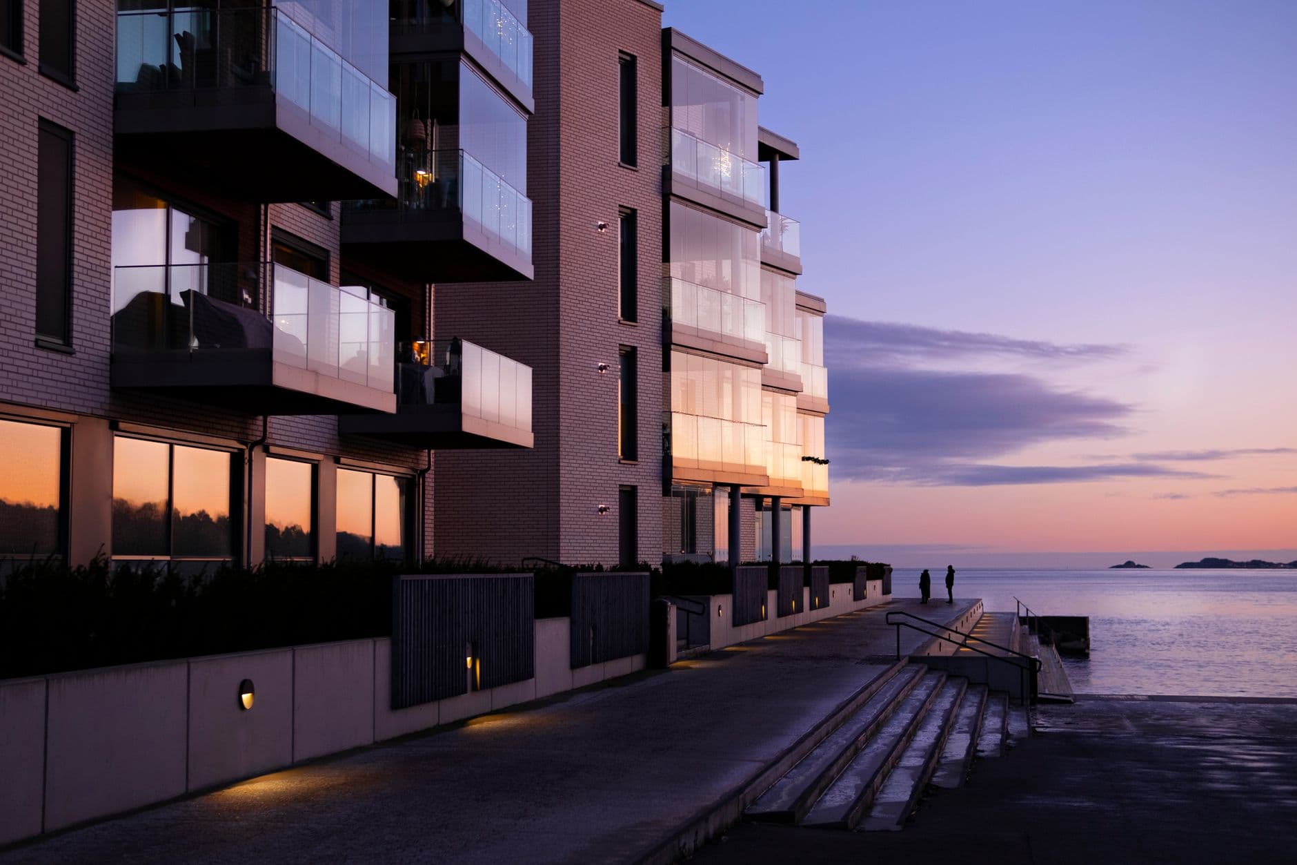 Moderne bakstenen appartementen met glazen balkons langs een promenade aan zee bij zonsondergang