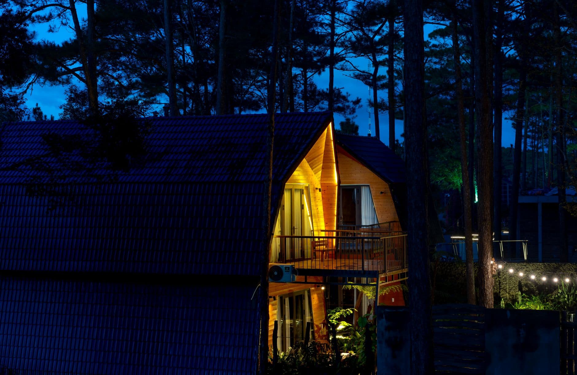 Houten chalet met verlichte ramen en balkon tussen hoge dennen in de avondschemering