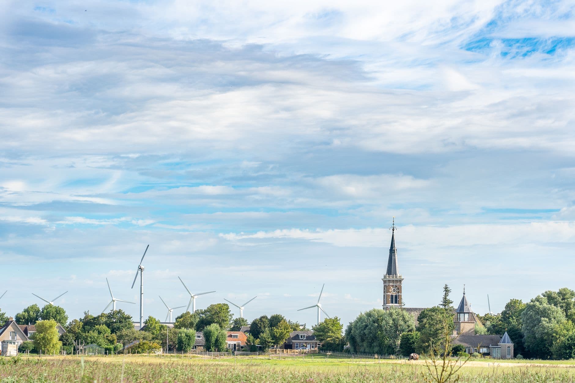 Dorpskerk met spitse toren tussen de bomen, met windturbines aan de rand van een veld