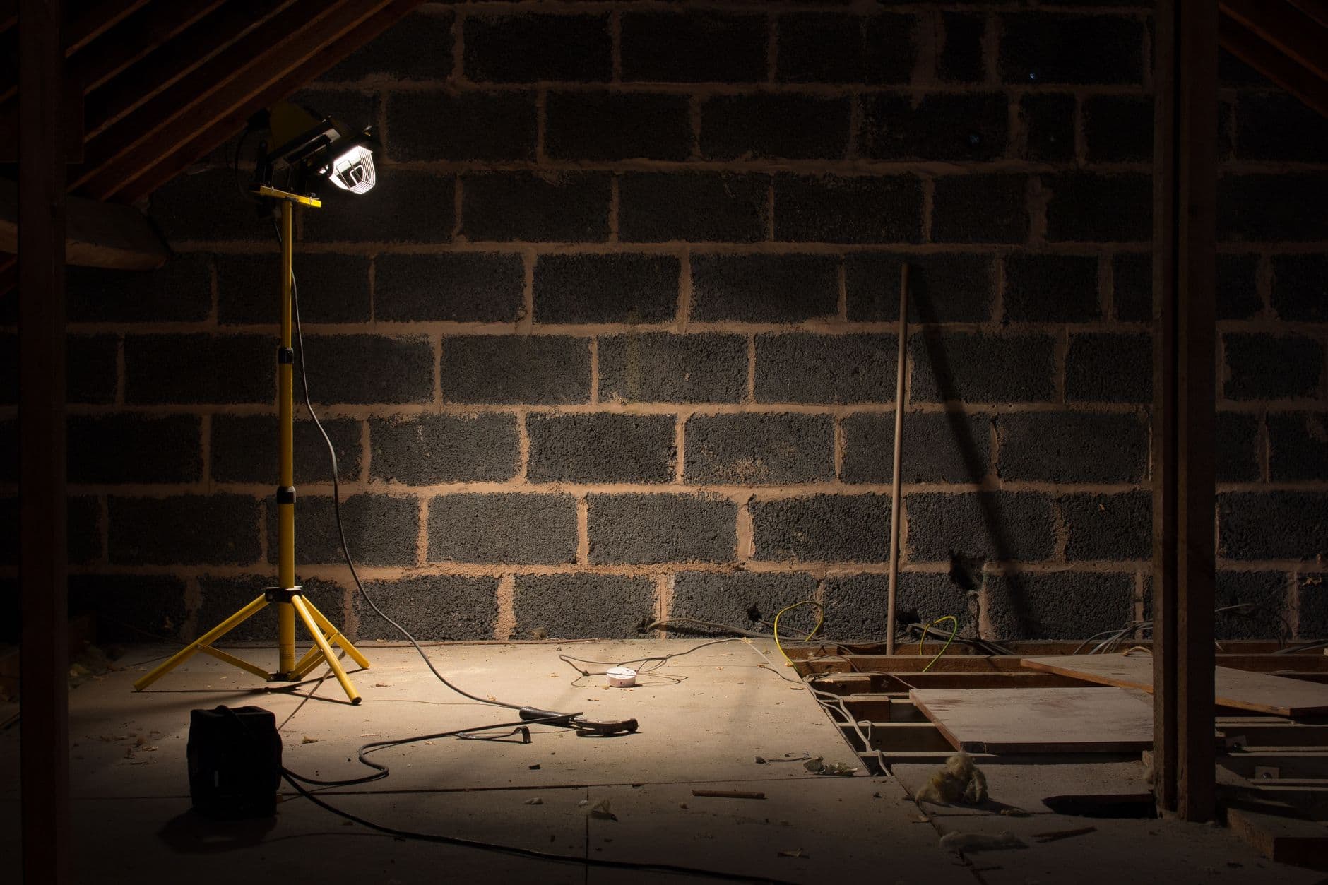 Zolder in renovatie met een bouwlamp op statief, losse kabels en een muur van betonblokken
