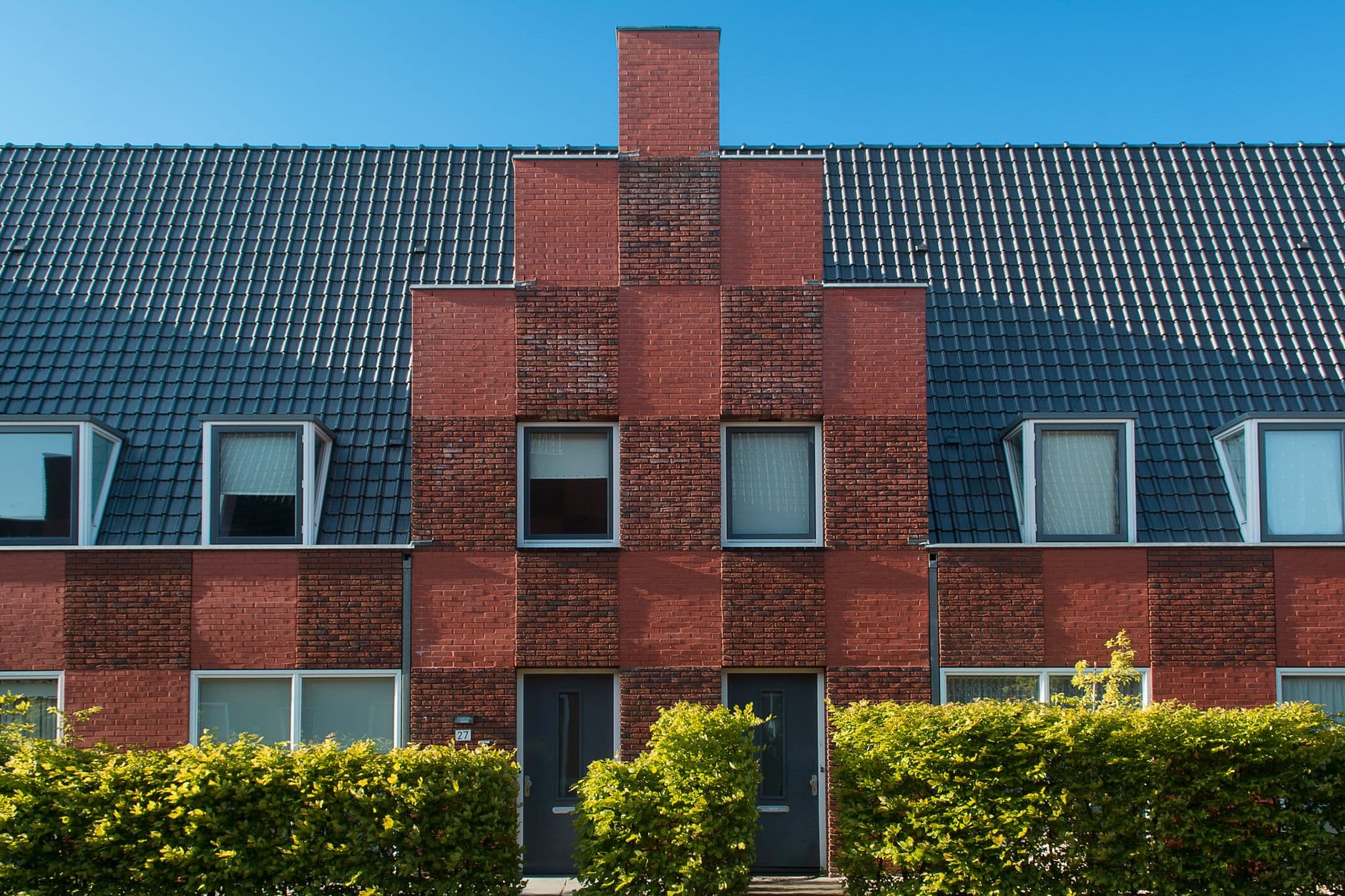 Rij bakstenen rijwoningen met donkere pannendaken, schoorsteen en groene hagen onder blauwe lucht