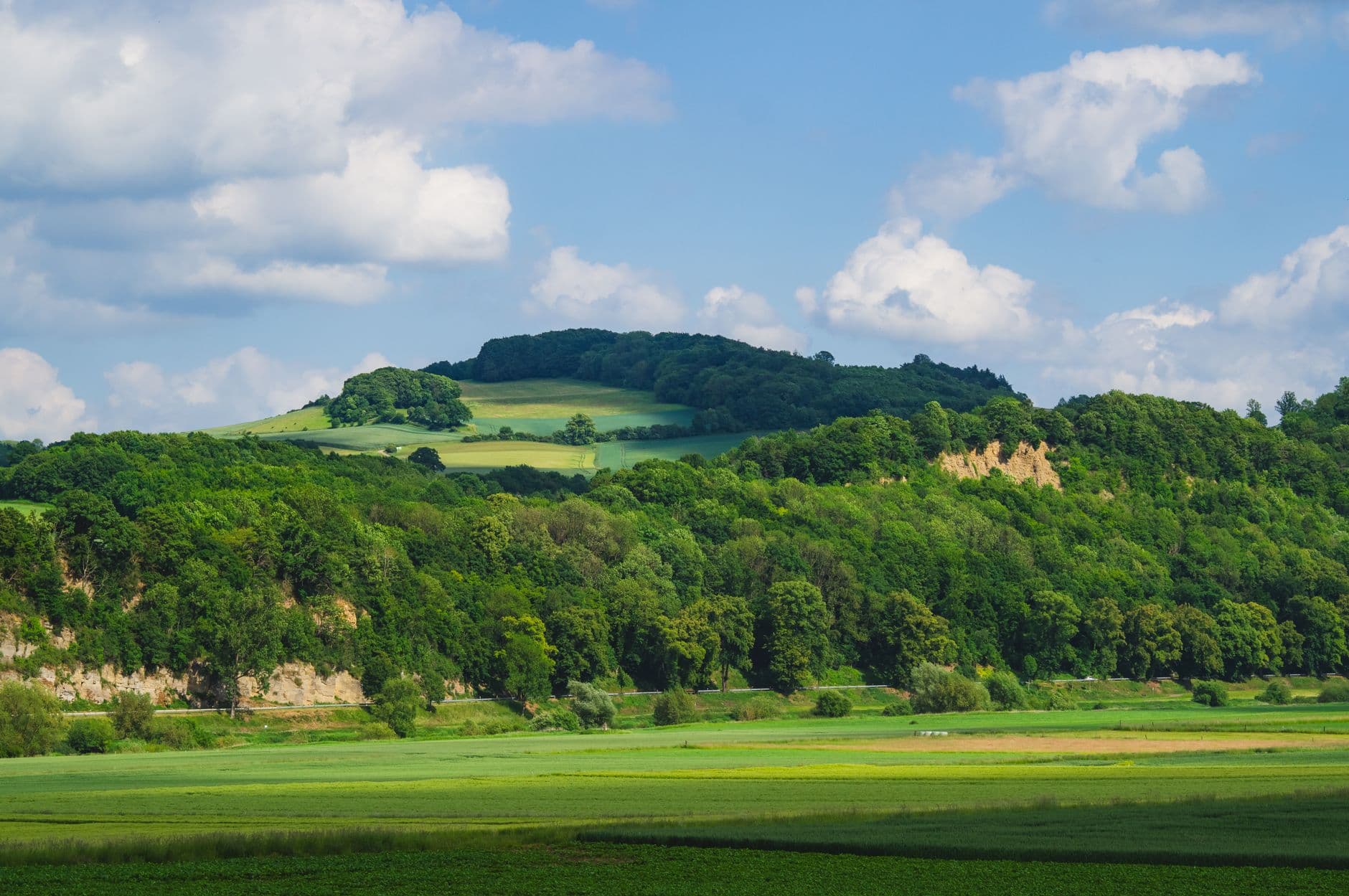 Glooiend groen landschap met akkers, beboste heuvels en stapelwolken aan een blauwe lucht