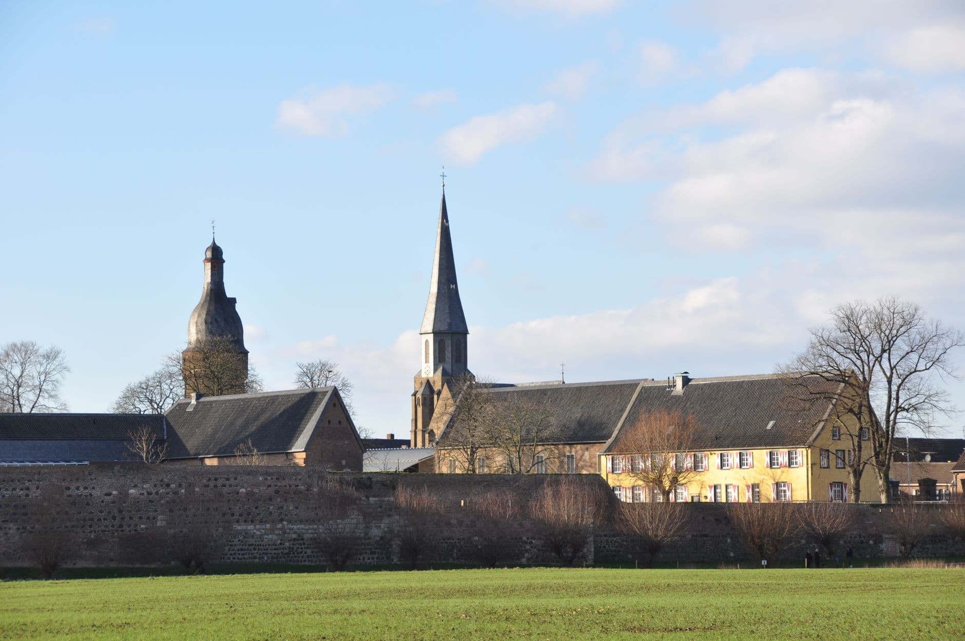 Dorpssilhouet met twee kerktorens en een oude muur achter een vlakke groene weide