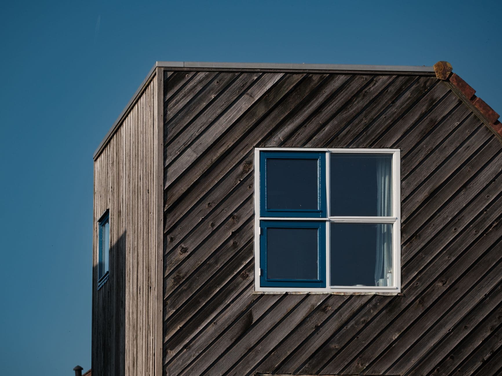 Gevel met donkere houten beplanking en een raam met blauwe kozijnen onder een blauwe lucht