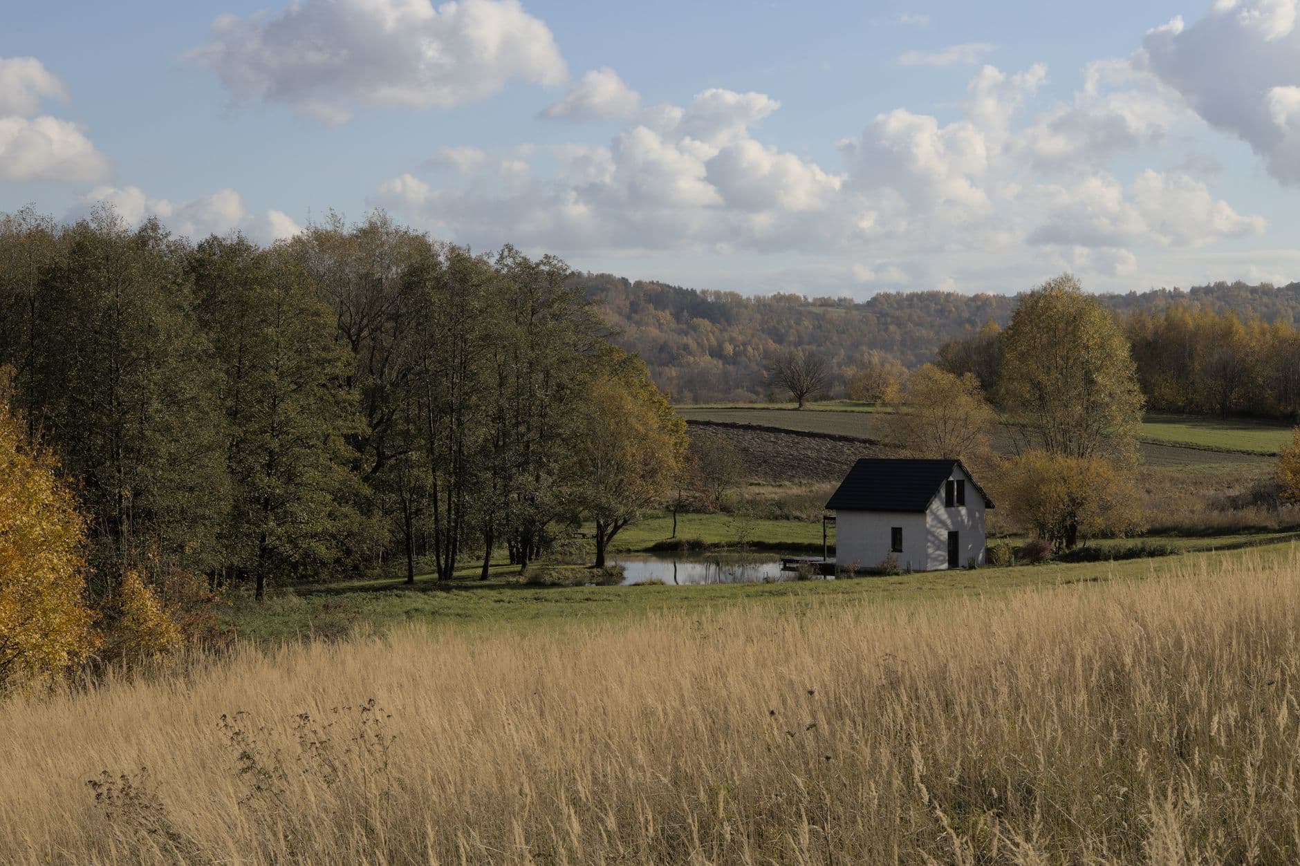 Landelijk tafereel met een wit huisje, een vijver en herfstbomen achter een veld met hoog gras