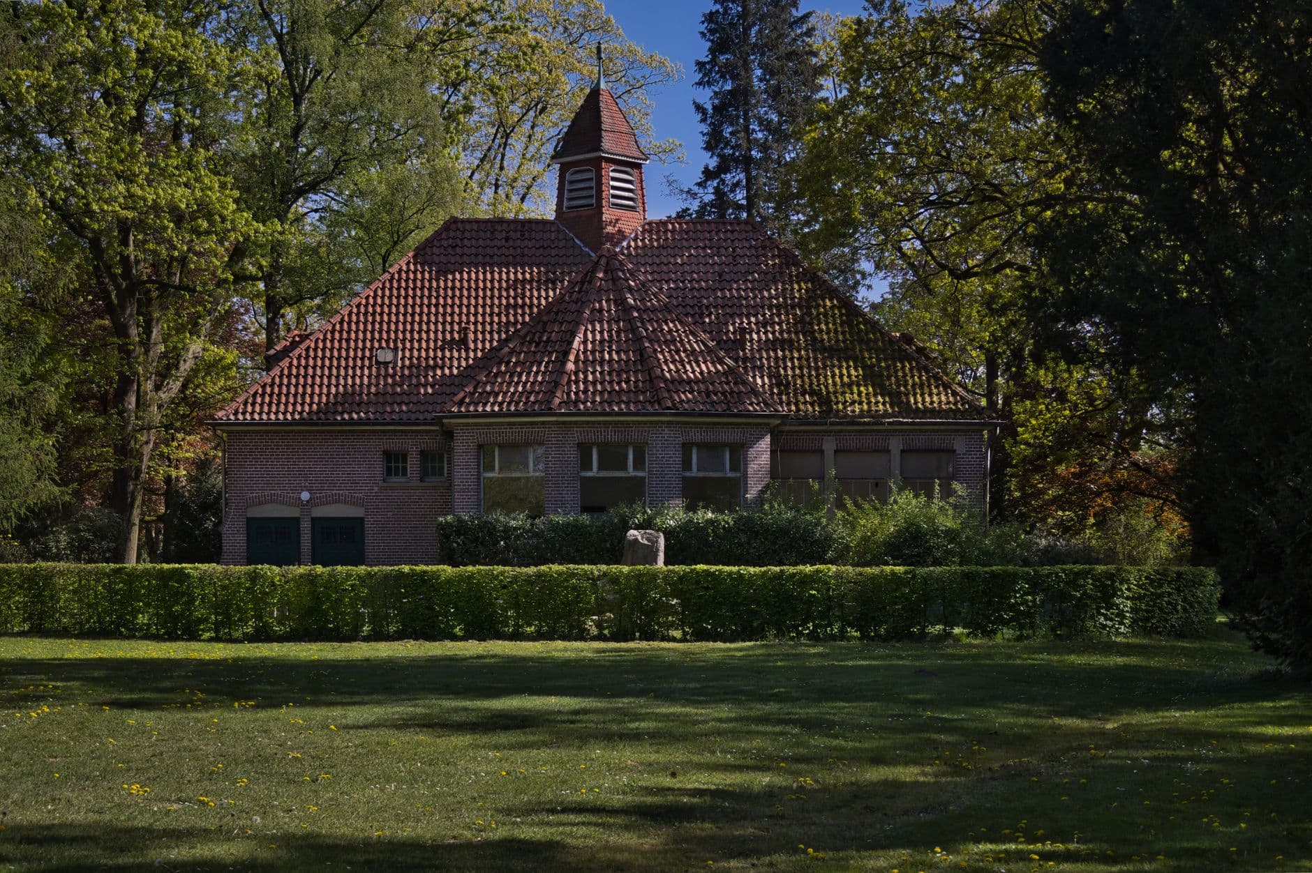 Bakstenen villa met rood pannendak en torentje, omringd door hoge bomen en een geschoren haag