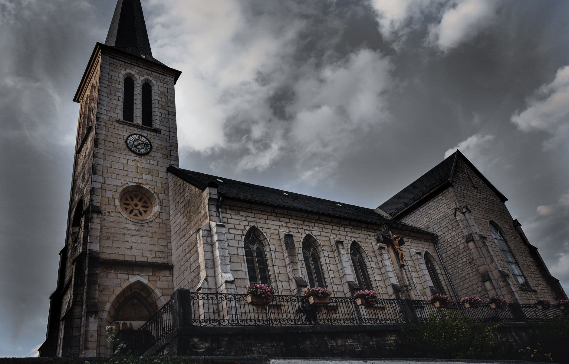 Neogotische stenen dorpskerk met klokkentoren en bloembakken onder een donkere bewolkte lucht
