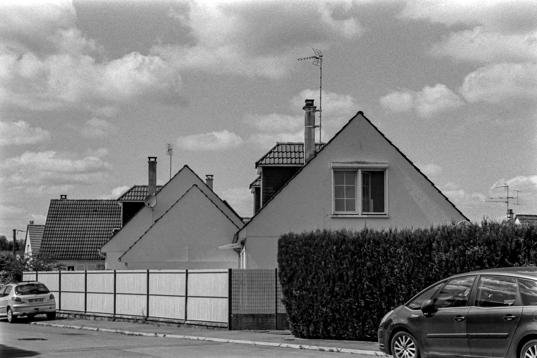 Zwart-witbeeld van woningen met puntgevels en pannendaken achter een haag en een tuinomheining