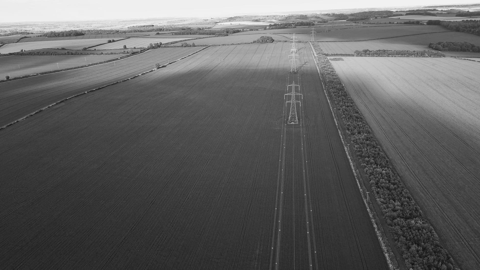 Luchtbeeld in zwart-wit van een hoogspanningslijn die recht over vlakke akkers loopt