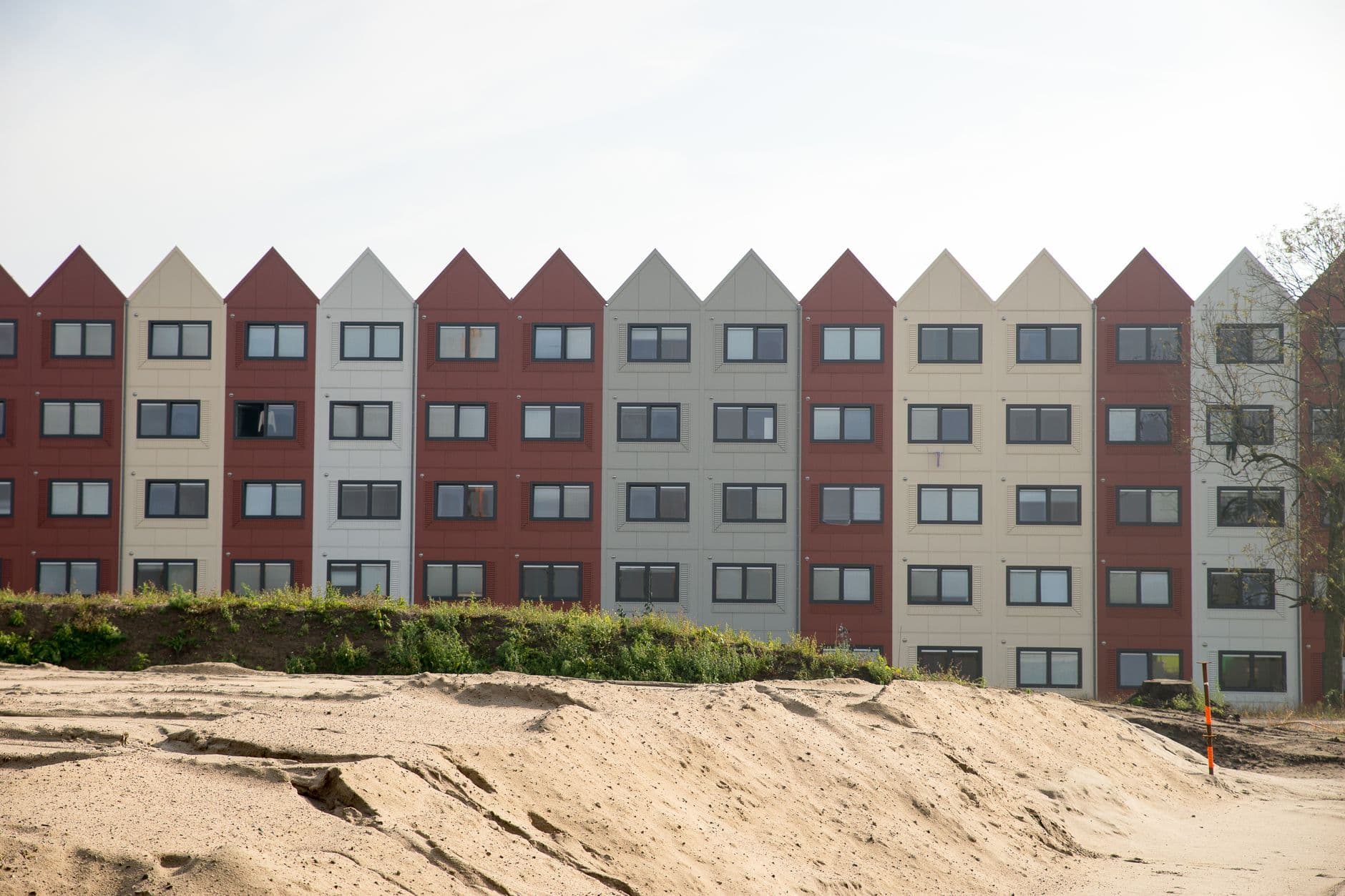 Nieuwbouwrij met puntgevels in rode, grijze en witte tinten achter een zandhoop op een bouwterrein