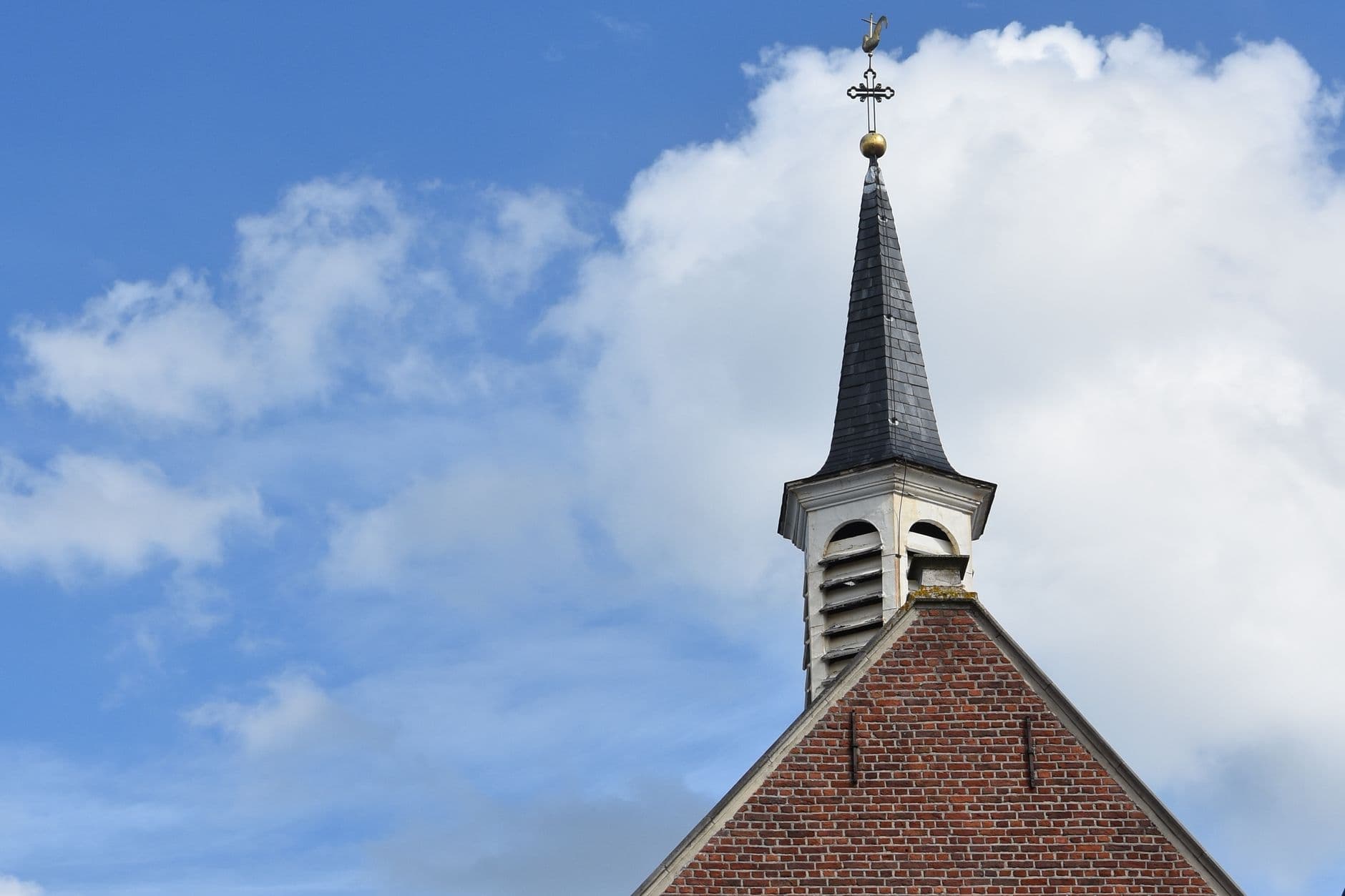 Bakstenen kapel met leien torenspits en kruis tegen een blauwe lucht met witte wolken