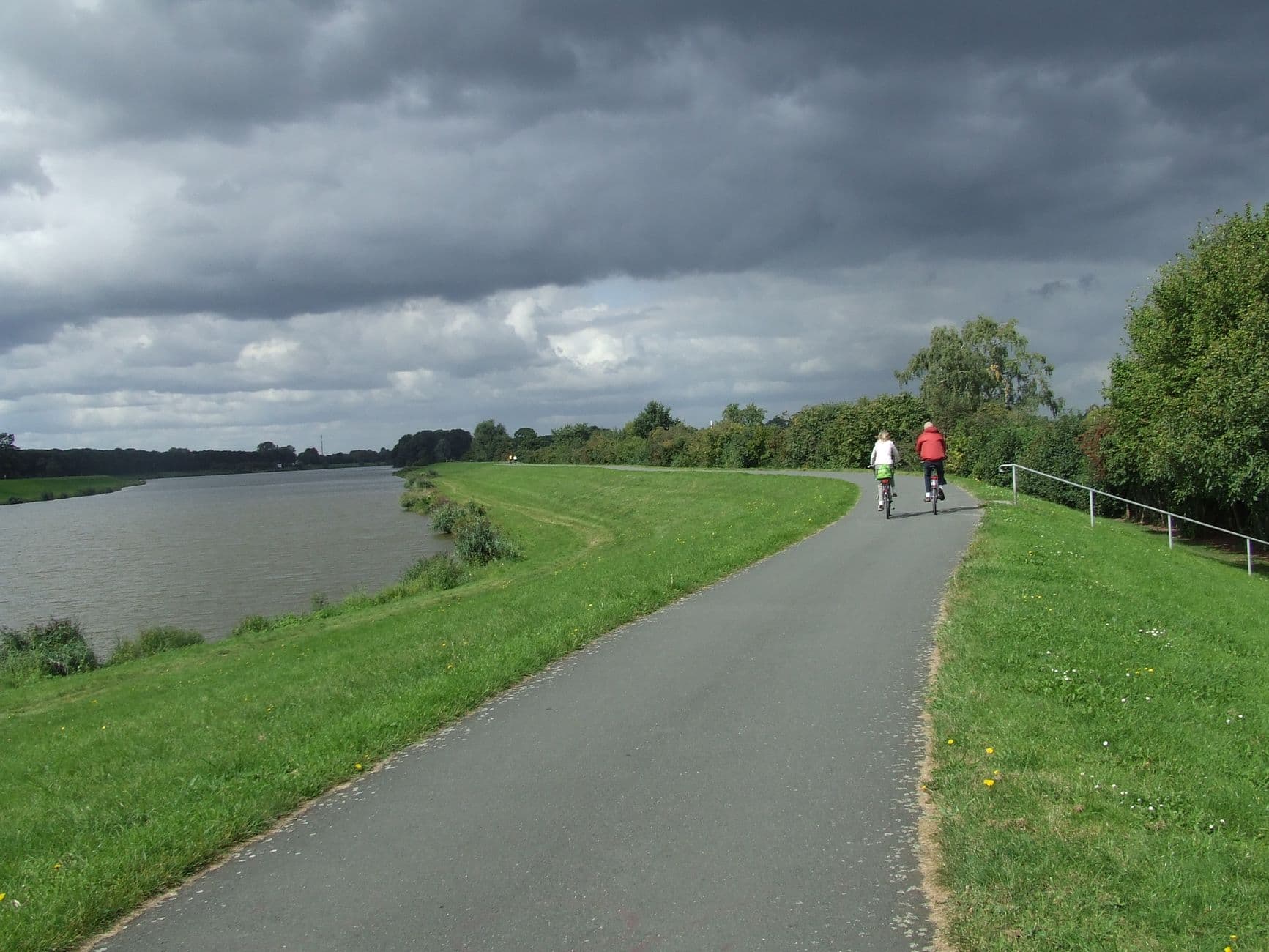 Fietspad op een dijk langs een rivier onder donkere wolken, met twee fietsers in de verte