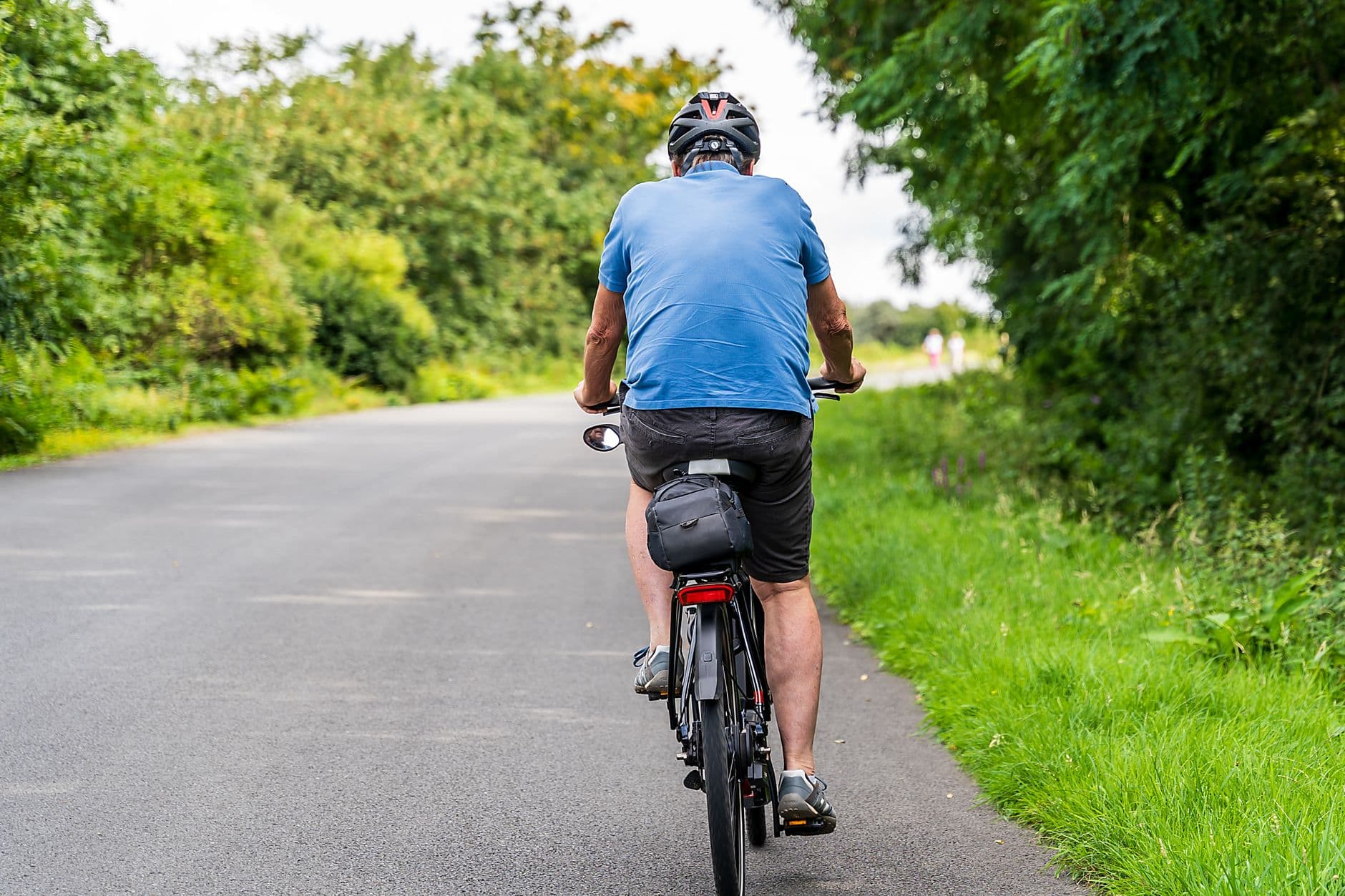 Oudere fietser met helm op een landelijke weg tussen groene hagen, gezien van achteren