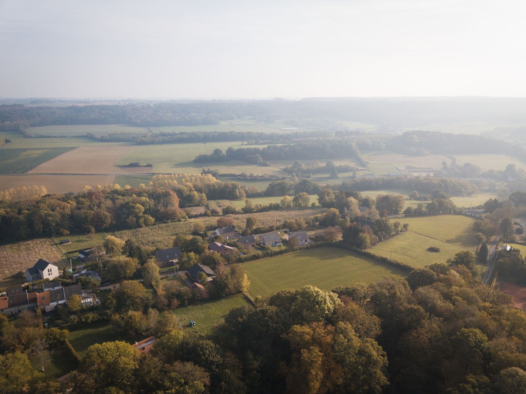 Luchtbeeld van een glooiend landschap met verspreide woningen, weiden en bossen in herfstkleuren