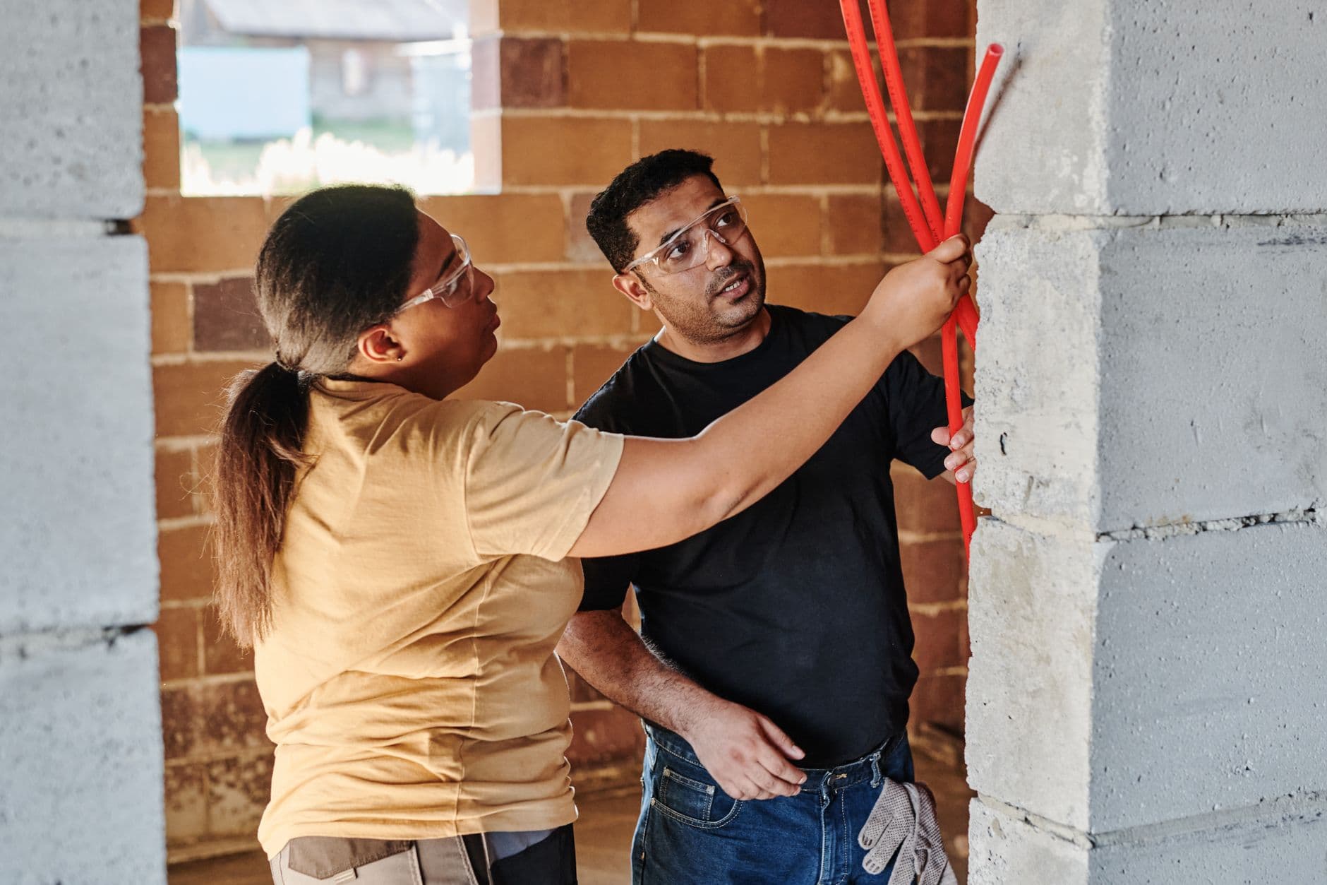 Two tradespeople inspecting red electrical conduits in a house under construction