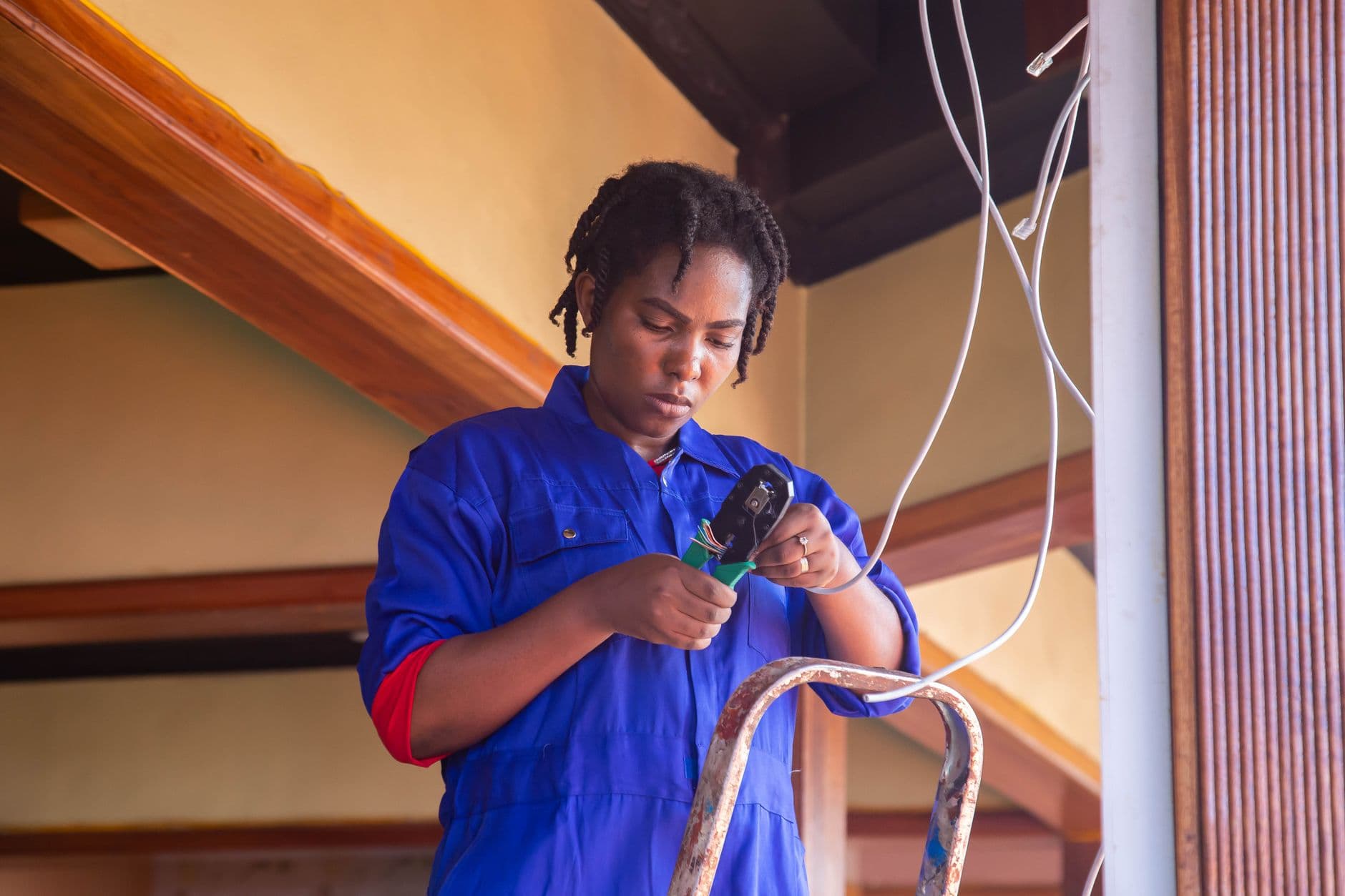 Elektricien in blauwe overall werkt met een tang aan een draad bovenaan een ladder